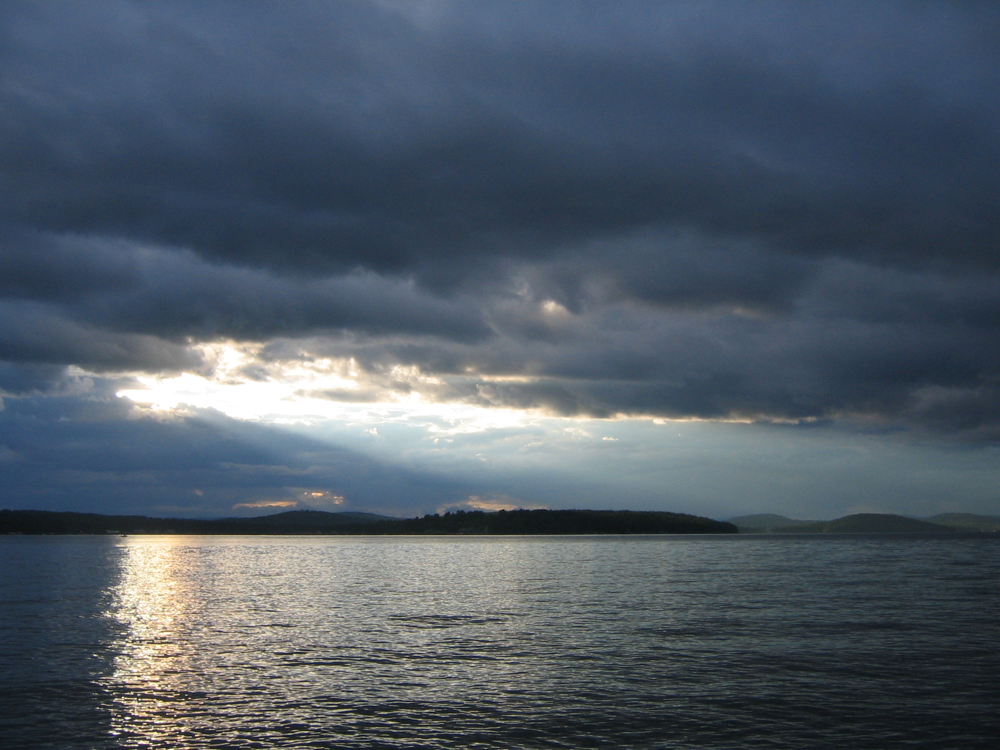 Uma vasta extensão de água sob um céu tempestuoso com raios de sol rompendo nuvens escuras, tons de azul e cinza, atmosfera contemplativa.